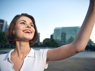 Fototapeta premium A cheerful young woman takes a selfie in a sunny urban park beside a river, smiling brightly as modern buildings rise in the background, conveying optimism, confidence, and a casual, upbeat lifestyle