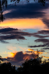 Vibrant sunset sky with dramatic dark clouds and a golden orange glow. Silhouetted tree branches frame the beautiful evening light