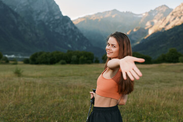 Obraz premium A cheerful young woman stands in a scenic valley, wearing a bright orange crop top. She smiles warmly and extends her arm toward the camera, creating a friendly, carefree outdoor mood.