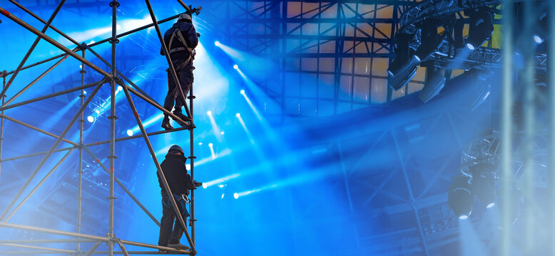 Stage workers in safety harnesses assemble scaffolding and adjust lighting equipment under bright beams in concert hall. Stage Equipment Installation.