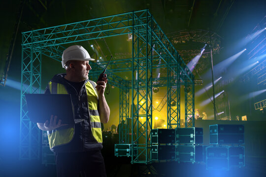 Engineer in hard hat and safety vest uses laptop and walkie-talkie while supervising concert stage light rig installation. Lighting Equipment Installation.