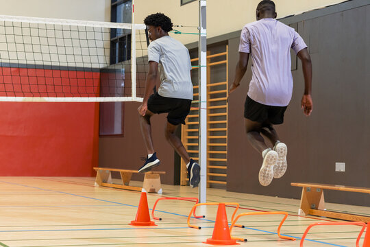 African american men jumping over hurdles at sports hall, volleyball net, cones
