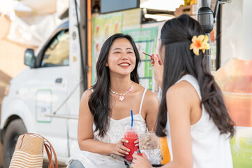 A friend putting on lipstick for pretty asian woman holding juice cup while sitting aside food truck