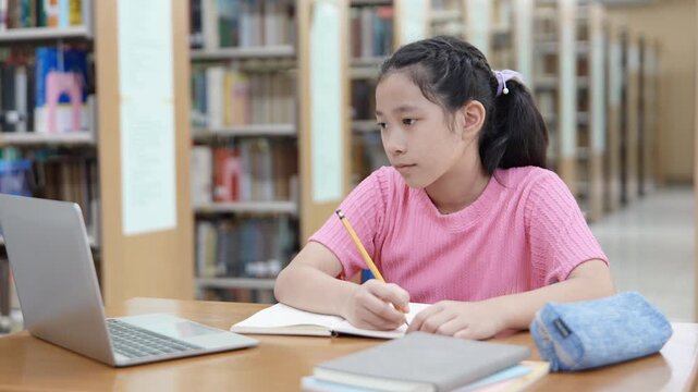 Focused girl doing homework and online learning at library using laptop. Concept of remote learning. Time management. Back to school. Education concept.