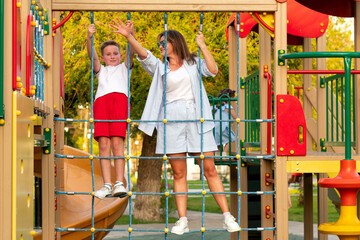Smiling mother and her son grip rope grid bridge at colorful park playground, balancing together and waving during lively summer bonding time outdoors