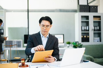 Obraz premium Professional Asian lawyer reading legal documents in modern office with gavel and scales on desk.