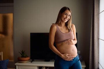 Happy mother to be day dreaming while holding her belly at home.