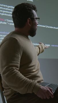 Medium vertical shot of Caucasian male coding teacher with laptop sitting on high chair, explaining computer script projected on screen, while conducting lecture in dark classroom