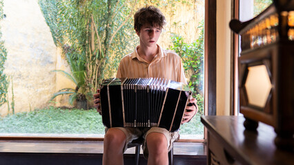 Teenager plays tango bandoneon inside a bright room with plants in the background