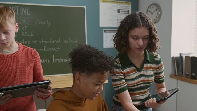 Medium pan shot of diverse children learning to write artificial intelligence generator software prompts on laptop and tablets at programming lesson at middle school, discussing tasks, giving advice