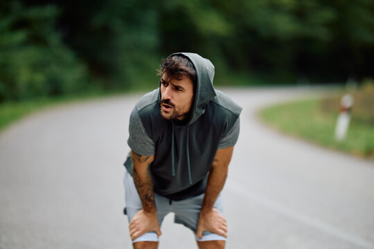 Athletic man taking a break from running, catching his breath on a curving road, feeling exhaustion after intense training