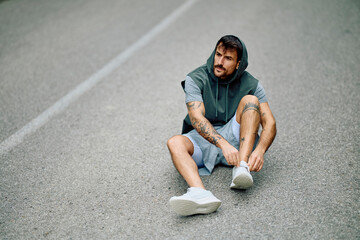 Young tattooed man in sportswear sitting on an asphalt road surface, tying his athletic shoes while...