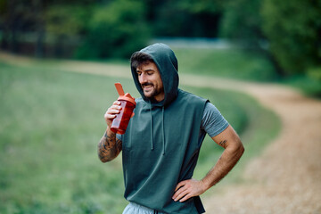 Man in a hoodie taking a hydration break from exercising outdoors, drinking from a sports shaker bottle