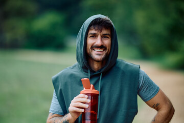 Young man smiling, wearing a hoodie, holding a water bottle after exercising outdoors. Healthy lifestyle and fitness