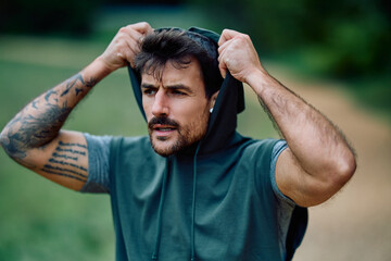 Athletic man with tattoos training outdoors. He is pulling up his hoodie, showing concentration and...
