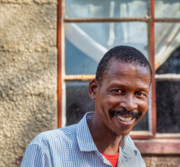 single african mature man portrait, in the village, standing in front of the house, toothy smile...