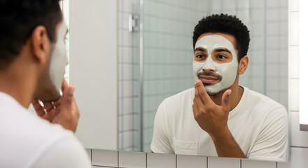 Young Black man applying a clay face mask while looking at his reflection in the bathroom mirror, focusing on skincare and grooming routine