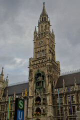 Munich's New Town Hall, a stunning building with its ornate Gothic facade, rises beneath a cloudy sky. The clock tower features an intricate Glockenspiel with moving figures.