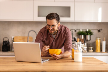 A man in a casual shirt eats cereal and uses a laptop in a stylish kitchen. He enjoys a balanced lifestyle, incorporating work, health, and relaxation into his daily routine.