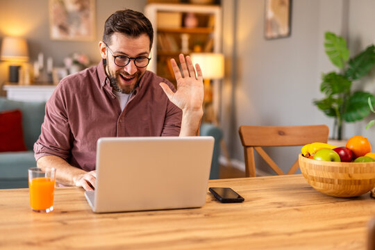 Friendly man greeting someone on a video call while working from home. He sits in a stylish kitchen with a laptop, orange juice, and warm lighting, creating a welcoming and professional setting.