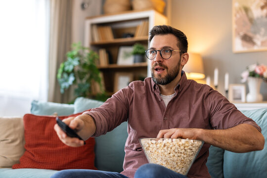 A man in a casual outfit sits on a stylish sofa, enjoying his favorite show. Holding a remote control and a bowl of popcorn, he appears relaxed and happy, creating a cozy home ambiance