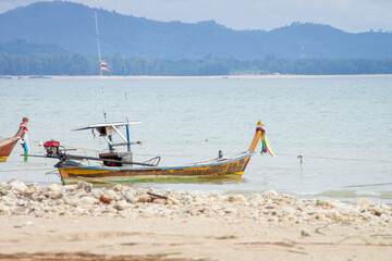 Naklejka premium Traditional Thai Fishing Boat Moored at a Rocky Tropical Shore. A colorful, classic longtail fishing boat anchored in the shallow water of a bay with rocky, sandy beach and distant forested mountains