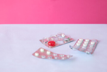 Three blister packs of pills on a white surface. One pack is partially empty, revealing a red capsule. The background is a solid pink color.