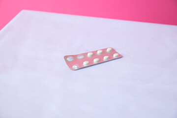A blister pack of white tablets on a light surface with a pink background. The pack contains several pills arranged in a grid pattern.