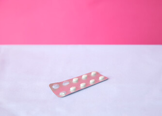 A blister pack of white tablets on a pink background. The pack is partially open, revealing several tablets. The scene is minimalistic and clean.