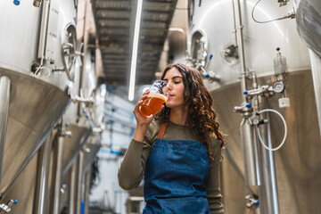 Female master brewer tasting freshly made beer from a glass, standing in a small craft brewery with stainless steel tanks