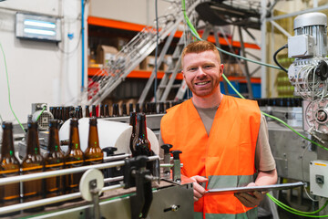 Smiling man examining beer bottle production with digital tablet in factory. Industry worker overseeing manufacturing