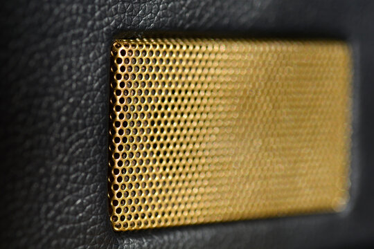 A close-up shot of a rectangular gold perforated speaker grill with shallow depth of field set against a black textured synthetic leather surface