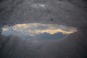 A puddle on a dirt surface reflects the sky with clouds at sunset. The scene captures the interplay of water and light in a natural setting.