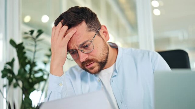 Worried businessman, dressed in a light blue shirt, processes upsetting news from a document in a bright office. Stressed specialist expresses frustration and disappointment with financial results. - Powered by Adobe