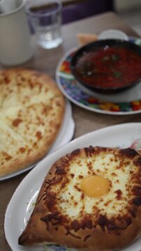 Vertical screen delicious Georgian cuisine with khachapuri adjaruli baked with egg and cheese khachapuri imeruli and bowl of red soup on wooden table in cozy restaurant atmosphere