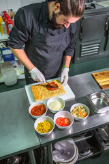 Professional male chef spreading tomato sauce over focaccia dough in commercial kitchen