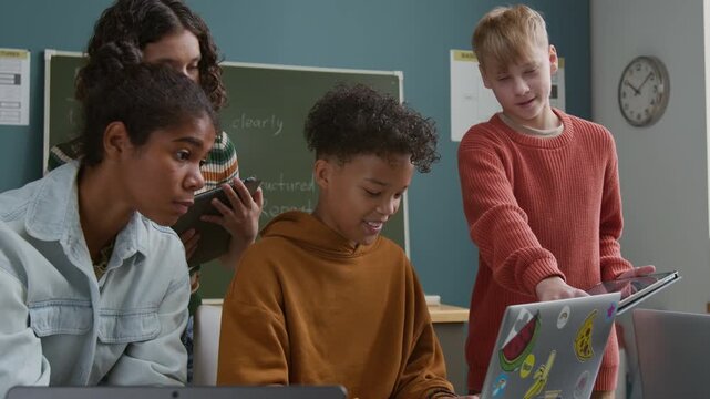 Medium shot of cheerful teenage African American boy sitting at desk in classroom at STEM lesson, writing prompt for artificial intelligence generative software, friends are watching and commenting