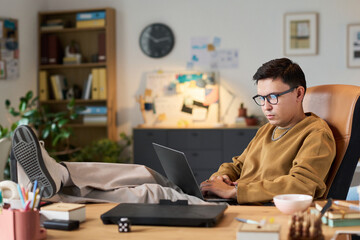 Asian man sitting at desk with legs up, working on laptop, wearing glasses, focused on screen,...