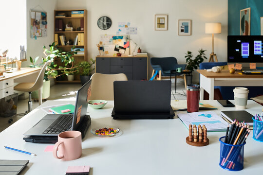 Modern office workspace featuring open desks with laptops, notebooks, stationery, coffee mug, candy, plants and digital devices arranged in collaborative environment, no people visible