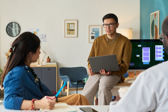 Asian man holding laptop leading discussion with Asian woman and Black man in modern office setting, digital project interface visible on screen in background