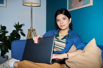 Portrait of Asian woman sitting on sofa holding laptop and looking into camera, relaxed posture, casual clothing, indoor setting with plant and framed artwork visible
