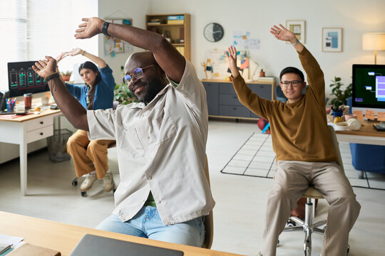 Diverse group of men and woman stretching arms while sitting in modern office, smiling and participating in workplace wellness activity during break, computer monitors in background