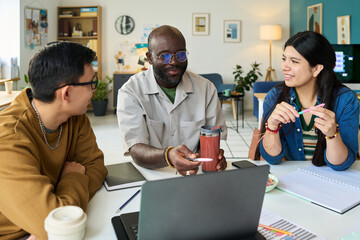 Black man explaining concept to Asian man and Hispanic woman while sitting at table with laptop and notebooks during collaborative meeting