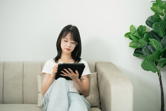 Young woman in casual outfit sitting on couch using tablet with focused expression on her face in modern living room with green plant - Powered by Adobe