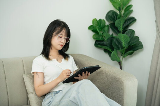 Young woman using digital tablet while sitting on modern couch in cozy living room surrounded by greenery and relaxed atmosphere