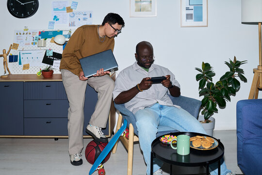 Man standing holding laptop talking to seated Black Man playing on smartphone in modern office setting with snacks and skateboard visible