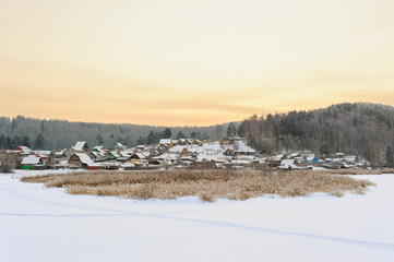Fototapeta premium A cozy winter village nestled beyond snowy fields, with frost-covered vegetation in the foreground. The forest rises behind the village, illuminated by the warm golden light of sunset, creating a peac