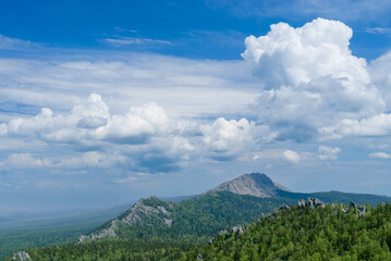 A stunning view of Taganay National Park featuring a lush forest with rocky outcrops in the foreground. Beyond the trees, a scenic mountain ridge stretches into the distance under a deep blue sky fill