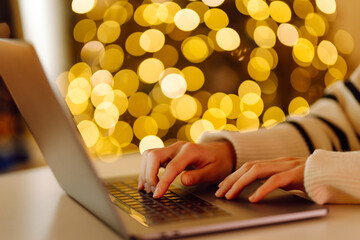 Close-up of hands typing on a laptop while sitting at a table in a cozy room. The hands of a young female freelancer working against backdrop of bright lights in room. Freelancing concept, keyboards.
