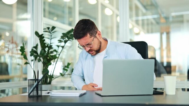 Suffering businessman in light blue shirt and white t-shirt experiences intense stomach pain at modern office desk. Male expresses severe discomfort and agony, interrupting work.